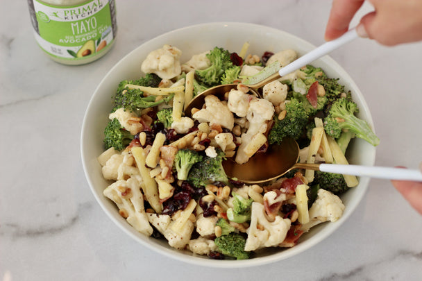 Hands using white and gold serving spoons in a bowl of broccoli cauliflower salad, with a jar of Primal Kitchen Mayo in the background. 