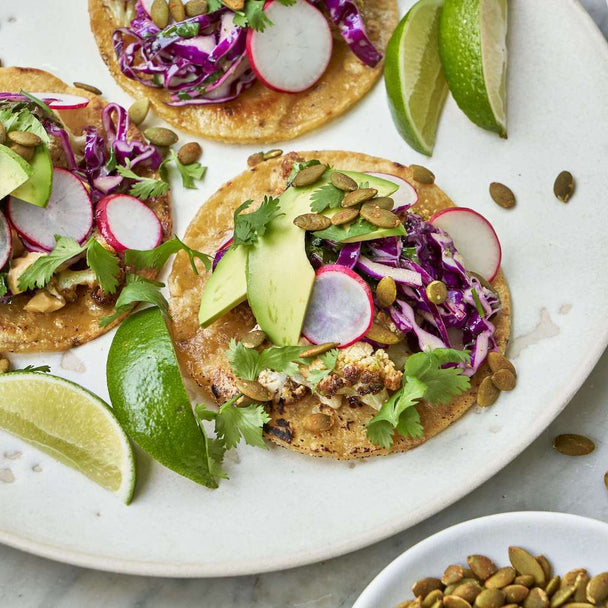 Three caramelized cauliflower tacos made with Primal Kitchen Cilantro Lime Dressing, on a white plate. Bowls of Three caramelized cauliflower tacos made with Primal Kitchen Chipotle Lime Mayo and pepitas beside the plate. 