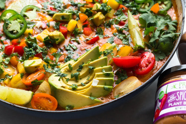  A skillet of chicken con queso topped with veggies and avocado slices, with a jar of Primal Kitchen Queso Dip in the background. 