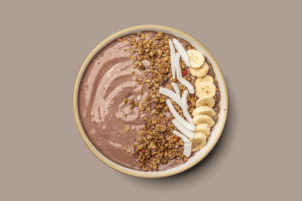 Overhead shot of a chocolate collagen smoothie bowl topped with granola, coconut, and banana slices, on a brown background. 