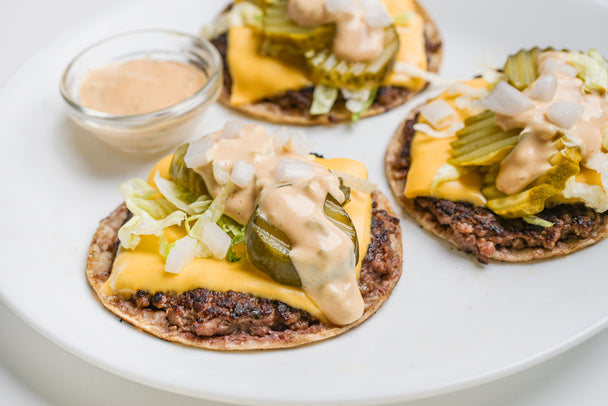 Three delicious cheeseburger patties on corn tortillas with a side of sauce. 