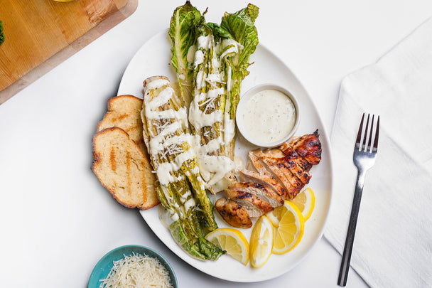 A plate of grilled romaine salad with Primal Kitchen Ranch next to a grilled chicken breast and toast.