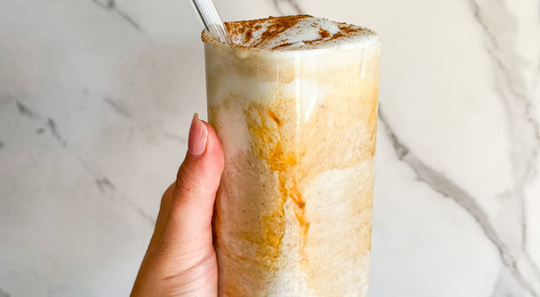 A woman’s hand holding a horchata smoothie in a clear glass, with a white marbled background. 