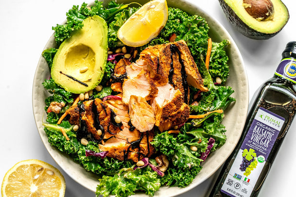 A bowl of kale salad with grilled salmon on top, next to a bottle of Primal Kitchen Balsamic Vinegar.