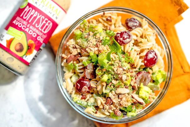 A clear glass bowl of poppyseed pasta salad, on an orange napkin, with a bottle of Primal Kitchen Poppyseed Dressing on the side. 