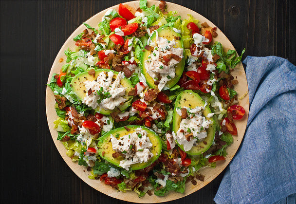 BLT chicken salad with Primal Kitchen Mayo inside of 4 avocado halves, on a plate. A blue napkin in the background. 