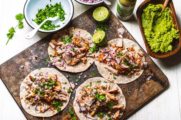 Four pork carnitas tacos on a serving board surrounded by bowls of avocado and herbs.  