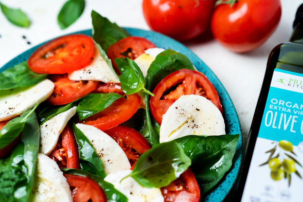 Closeup on a caprese salad with tomatoes and mozzarella on a blue plate, next to a bottle of Primal Kitchen Olive Oil. 