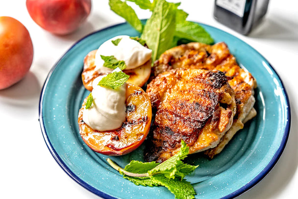 Several grilled chicken thighs with peaches and balsamic, on a blue plate. Peaches and balsamic vinegar bottle in the background. 