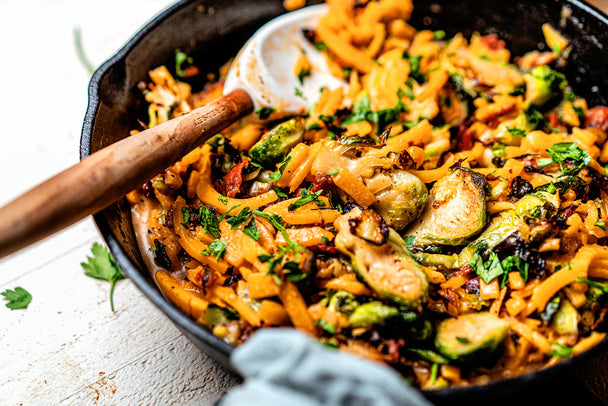 Spooning into a skillet of butternut squash pasta with bacon and Brussels sprouts made with Primal Kitchen No-Dairy Alfredo Sauce. 