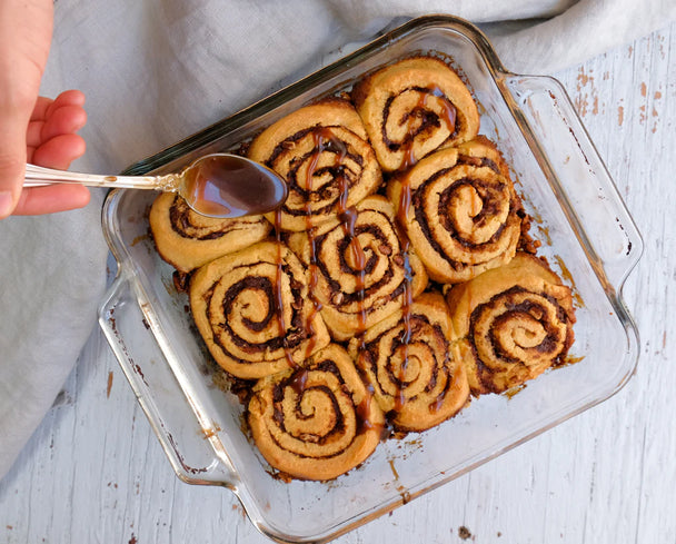 A hand with spoon drizzling caramel over a tray of pecan sticky buns.