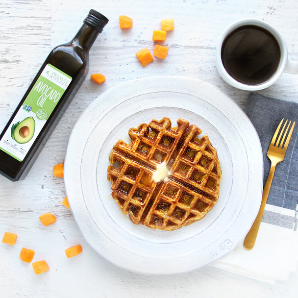 A butternut squash waffle on a white plate with a bottle of Primal Kitchen Avocado Oil, fork, napkin, and cup of coffee in the background.