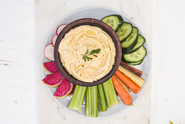 A wooden bowl of hummus surrounded by cut veggies, on a marble cutting board.