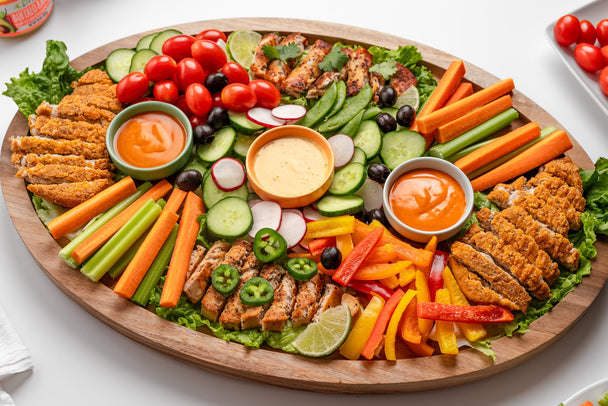 An oval-shaped wooden snack board piled with cut veggies, sliced mock chicken cutlets, and ramekins of Primal Kitchen Buffalo sauces & dips.  