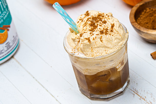 A clear glass of pumpkin whipped iced coffee with a blue straw. A bowl of cinnamon and a canister of collagen in the background.