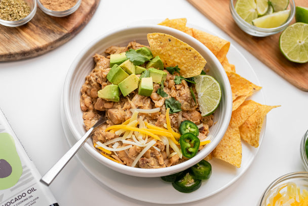 A bowl of white chicken chili on a white plate with a side of tortilla chips. Cutting boards and bowls with various ingredients in the background. 