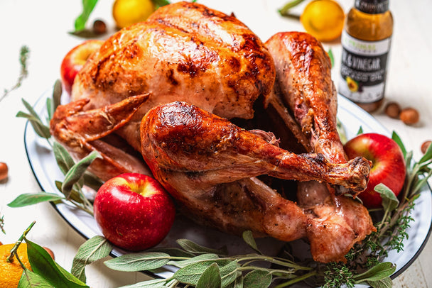 A juicy oven-baked whole turkey on a white platter surrounded by apples, herbs, and a bottle of Primal Kitchen Oil & Vinegar.