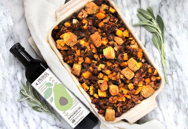 A white baking dish filled with stuffing, next to a bottle of Primal Kitchen Avocado Oil, a linen napkin, and a fresh herb.