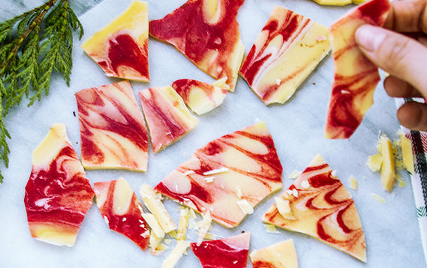 Pieces of white chocolate peppermint bark on a marble slab, with a sprig of greenery and a hand holding one piece of candy bark. 