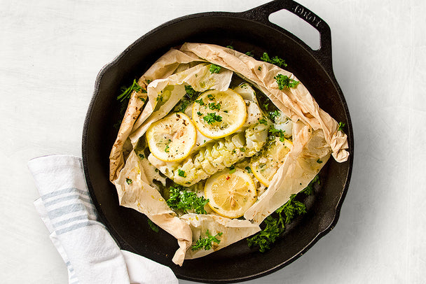 A parchment paper packet of white fish with lemon and herbs, inside a cast iron skillet. 