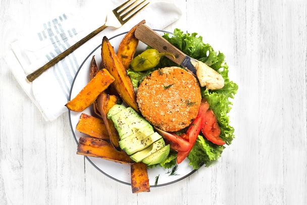 A salmon cake on a white plate surrounded by fries, sliced avocado, greens, and pickles.