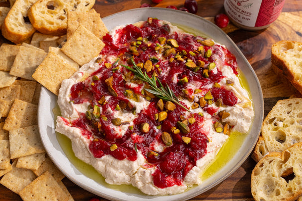  A plate of cranberry whipped feta dip and crackers on a beige platter, surrounded by bread and crackers for dipping.