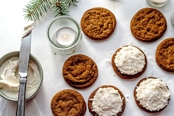  Gingerbread cookies, some frosted and some plain, on white parchment paper. A candle, greenery, and a jar of frosting with a knife in the background.