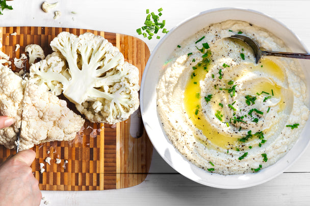 A bowl of ranch mashed cauliflower next to a cutting board with a hand chopping cauliflower.