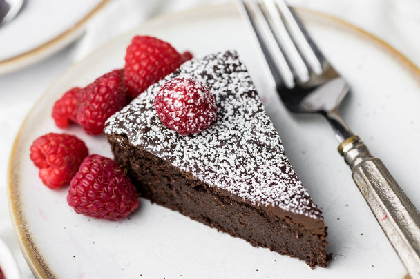 A slice of rich flourless chocolate cake dusted with powdered sugar and fresh raspberries on a china plate.