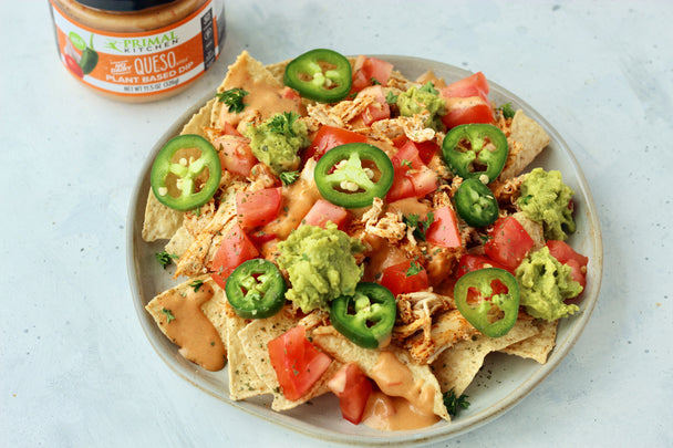 A plate full of chicken nachos with a jar of Primal Kitchen Queso Style Dip in the background.