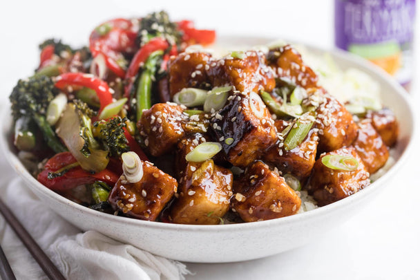 Teriyaki tofu and mixed vegetables atop cauliflower rice in a white bowl, with a bottle of Primal Kitchen No-Soy Teriyaki Sauce in the background. 