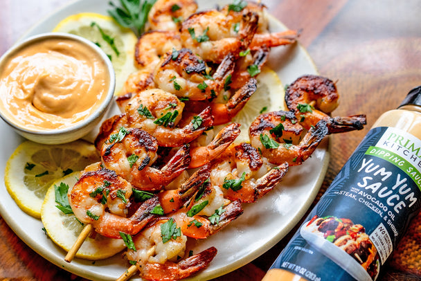 A plate of shrimp skewers with lemon slices and a bowl of dipping sauce next to a bottle of Primal Kitchen Yum Yum Sauce. 