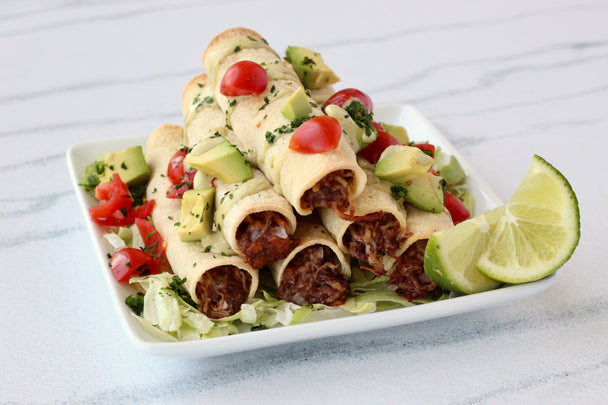 A pile of chicken taquitos on a white plate with lime slices, on a marble background.