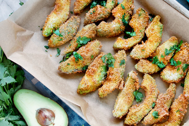 Breaded avocado fries on a baking sheet lined with parchment paper, next to half an avocado and fresh parsley.