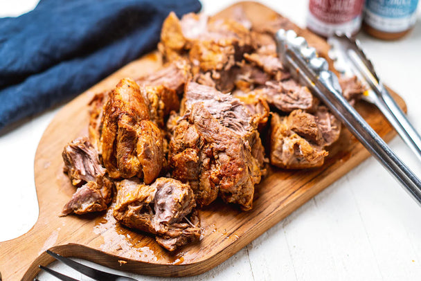 Cooked pulled pork on a wooden cutting board with silver tongs. Primal Kitchen Classic BBQ Sauce and Golden BBQ Sauce in the background.