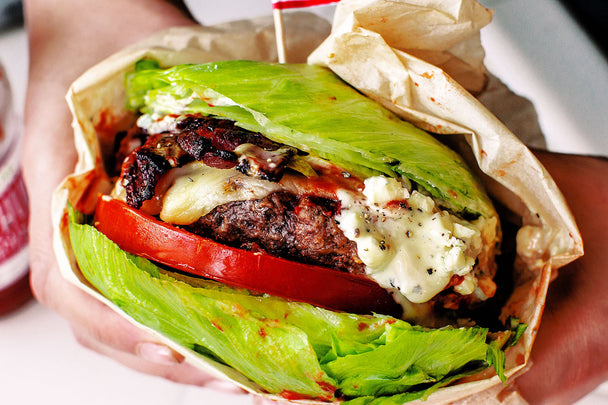 Hands holding a burger wrapped in lettuce and paper. The burger is topped with bacon, sliced tomato, and melty cheese. Primal Kitchen Ketchup in background.