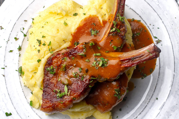 Overhead shot of two lamb chops with gravy atop mashed potatoes on a white plate.