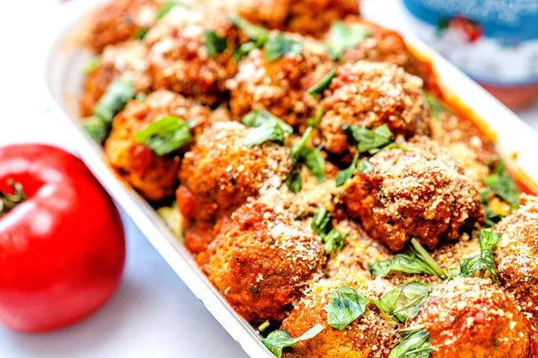 Tray of meatballs in marinara topped with fresh basil alongside a whole tomato and a jar of Primal Kitchen Roasted Garlic Marinara Sauce.