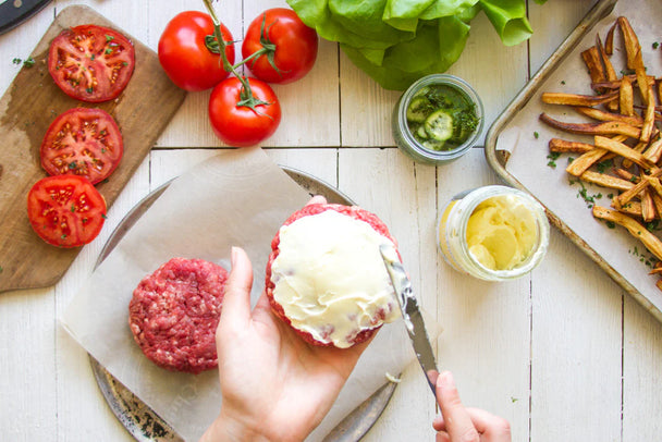 Overhead shot of hands spreading Primal Kitchen Garlic Aioli Mayo on a burger, fresh tomatoes, pickles, parsnip fries.