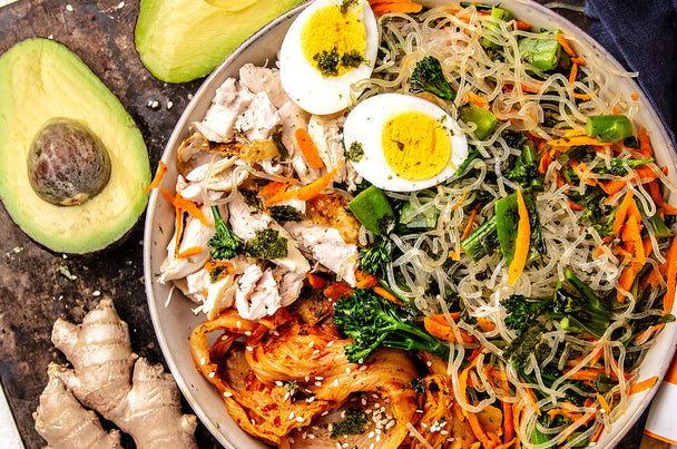 Shirataki noodle bowl with chicken, broccolini, shredded carrots, kimchi, and hard-boiled egg. Avocado and fresh ginger root on the side. 