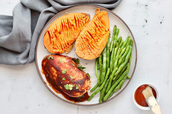 A plate of oven baked BBQ chicken, green beans, and sweet potatoes next to a cup of Primal Kitchen BBQ Sauce. 