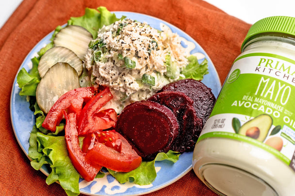 On a blue plate, a generous scoop of chicken salad on a bed of lettuce with beets, tomato slices, and pickle chips. An orange-red napkin and a jar of mayo in the background.