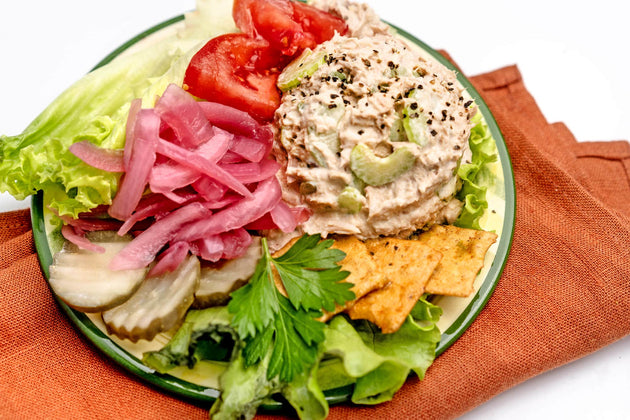 A scoop of tuna salad on a plate with pickles, onions, lettuce, tomato slices, and crackers, on a rust-colored napkin.