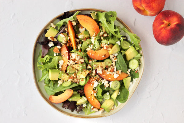  In a shallow bowl, a summer peach salad drizzled with Primal Kitchen Balsamic Dressing. A split peach in the foreground. 