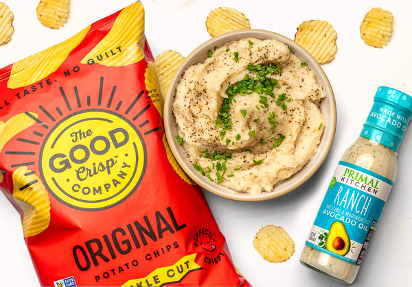 A bowl of potato chip mashed potatoes next to a bottle of Primal Kitchen Ranch dressing and a red bag of The Good Crisp Company potato chips. 