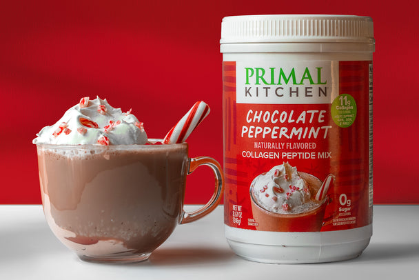 Peppermint hot chocolate garnished with candy cane pieces, in a clear glass mug. Next to it, a canister of Primal Kitchen Chocolate Peppermint Collagen. 