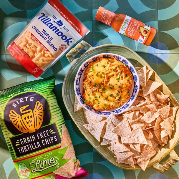 Easy Buffalo Chicken Dip in a bowl on a plate of chops, next to a bottle of Primal Kitchen Buffalo Sauce, a bag of Siete chips, and a bag of Tillamook cheese.