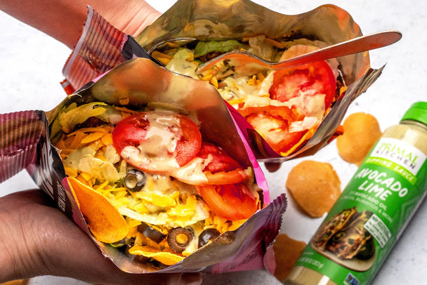 Overhead view into two bags of Wilde protein chips loaded with walking taco toppings, with a bottle of Primal Kitchen Avocado Lime Sauce in the background.