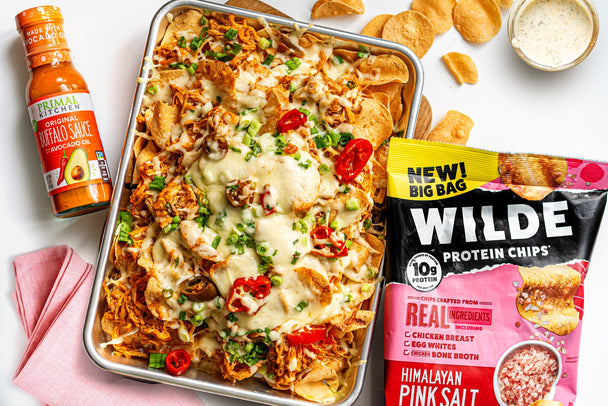 A tray of buffalo chicken nachos next to a bottle of Primal Kitchen Buffalo Sauce and a bag of Wilde protein chips.