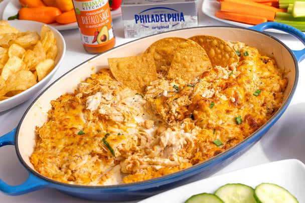 An oval dish of buffalo chicken dip with a few tortilla chips in it. A bottle of Primal Kitchen Buffalo Sauce and a block of Philadelphia Cream Cheese in the background with assorted chips and veggies.